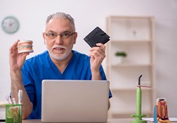 An older dentist holding mock dentures and a wallet
